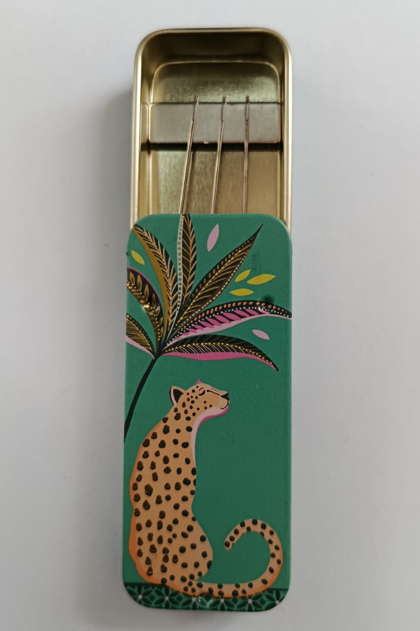 Small rectangular tin with a leopard and leaf design on a green background containing magnet to hold sewing needles in place