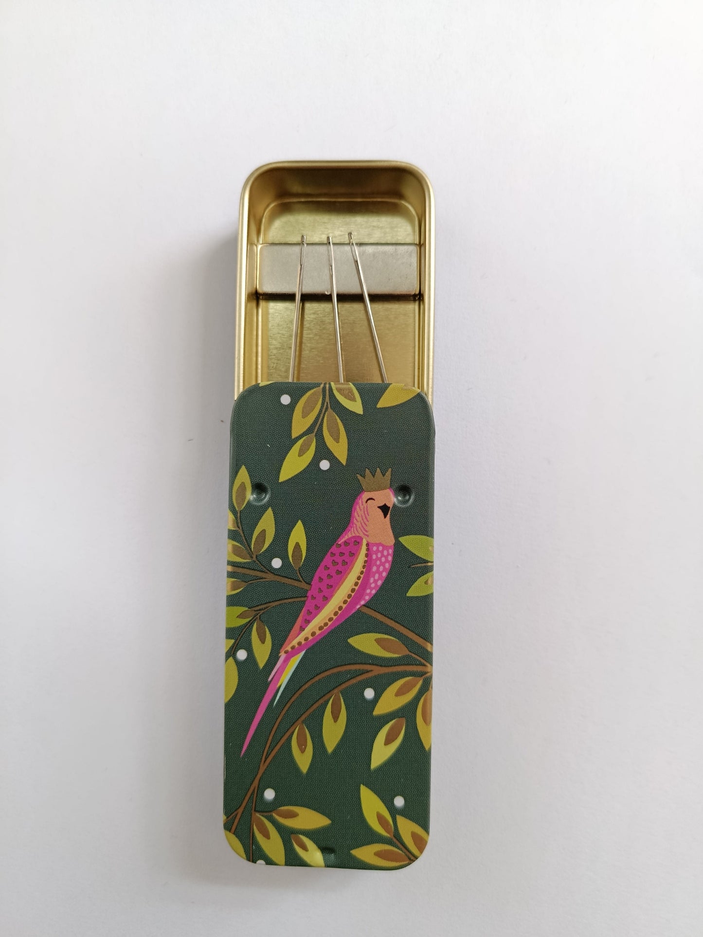Open slider tin containing magnet holding sewing needles in place. Pretty floral and parrot design on lid