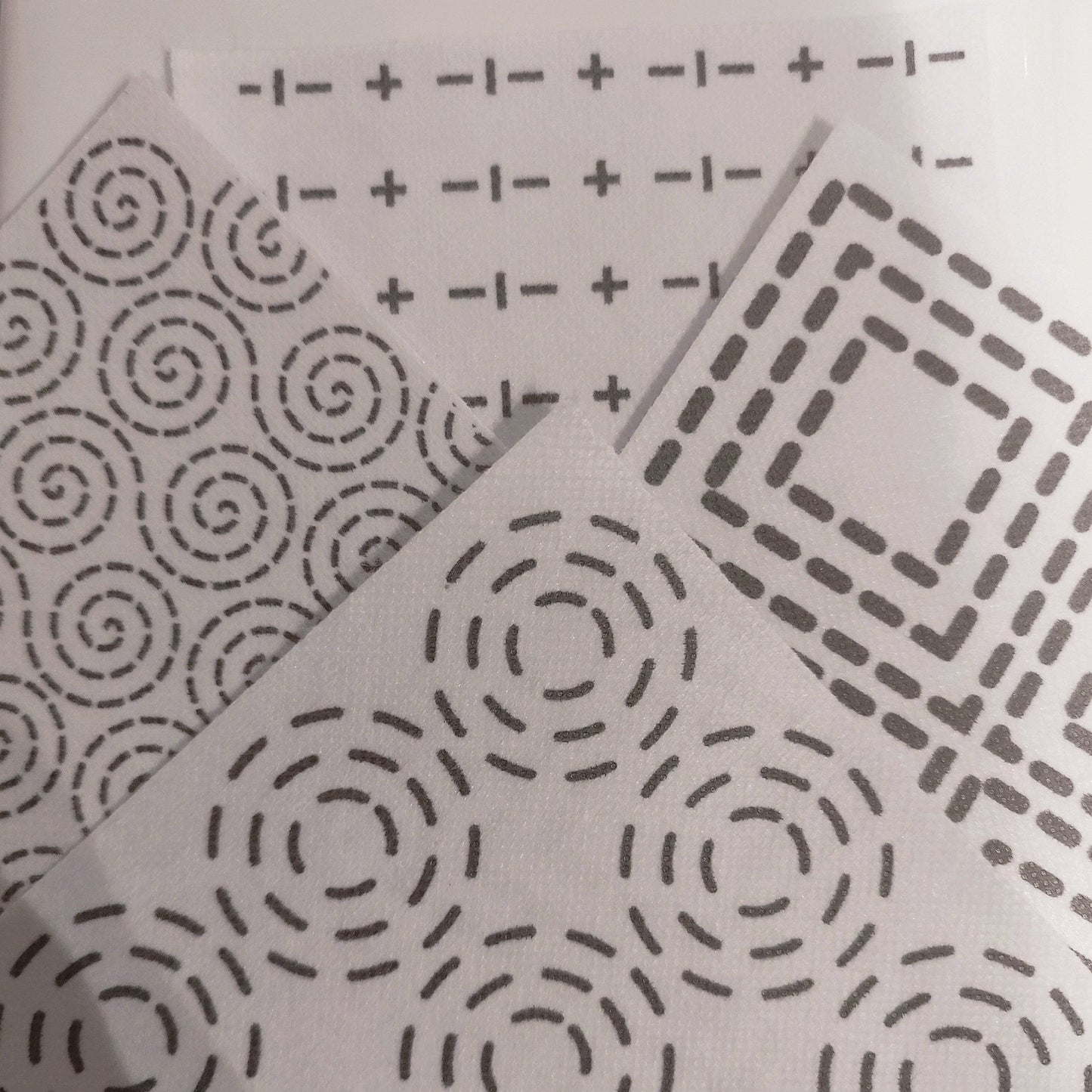 4 Sashiko Transfers. In the foreground is a concentric circles pattern. On the right is a concentric square pattern. At the back is a crosses and dashes sashiko pattern. On the left is a swirls dashed sashiko pattern