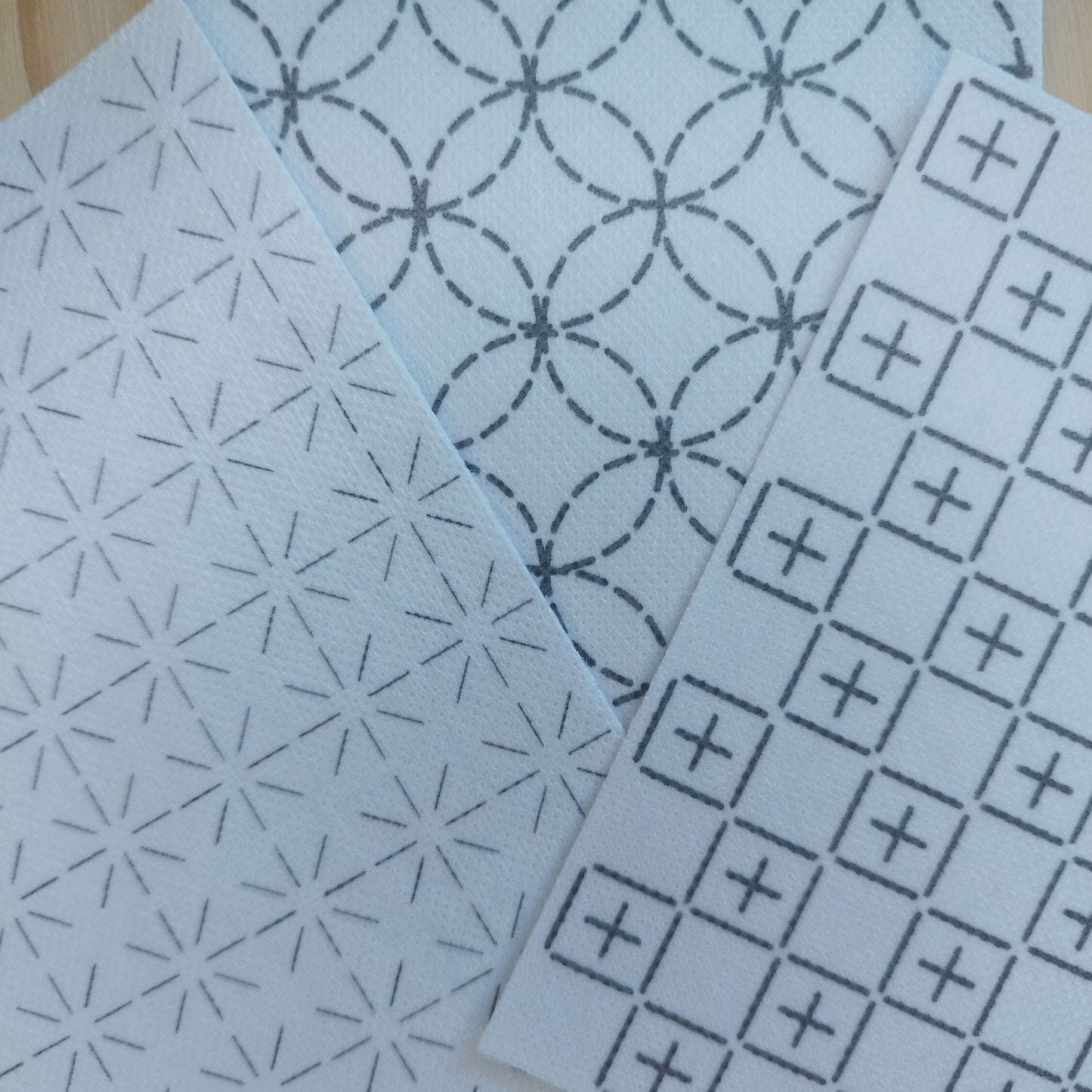 3 Sashiko Transfer Designs laid together on a table. The design on the right has crosses within squares. The design on the left is repeated 8 line crosses/daisys. The design at the back is overlapping circles.
