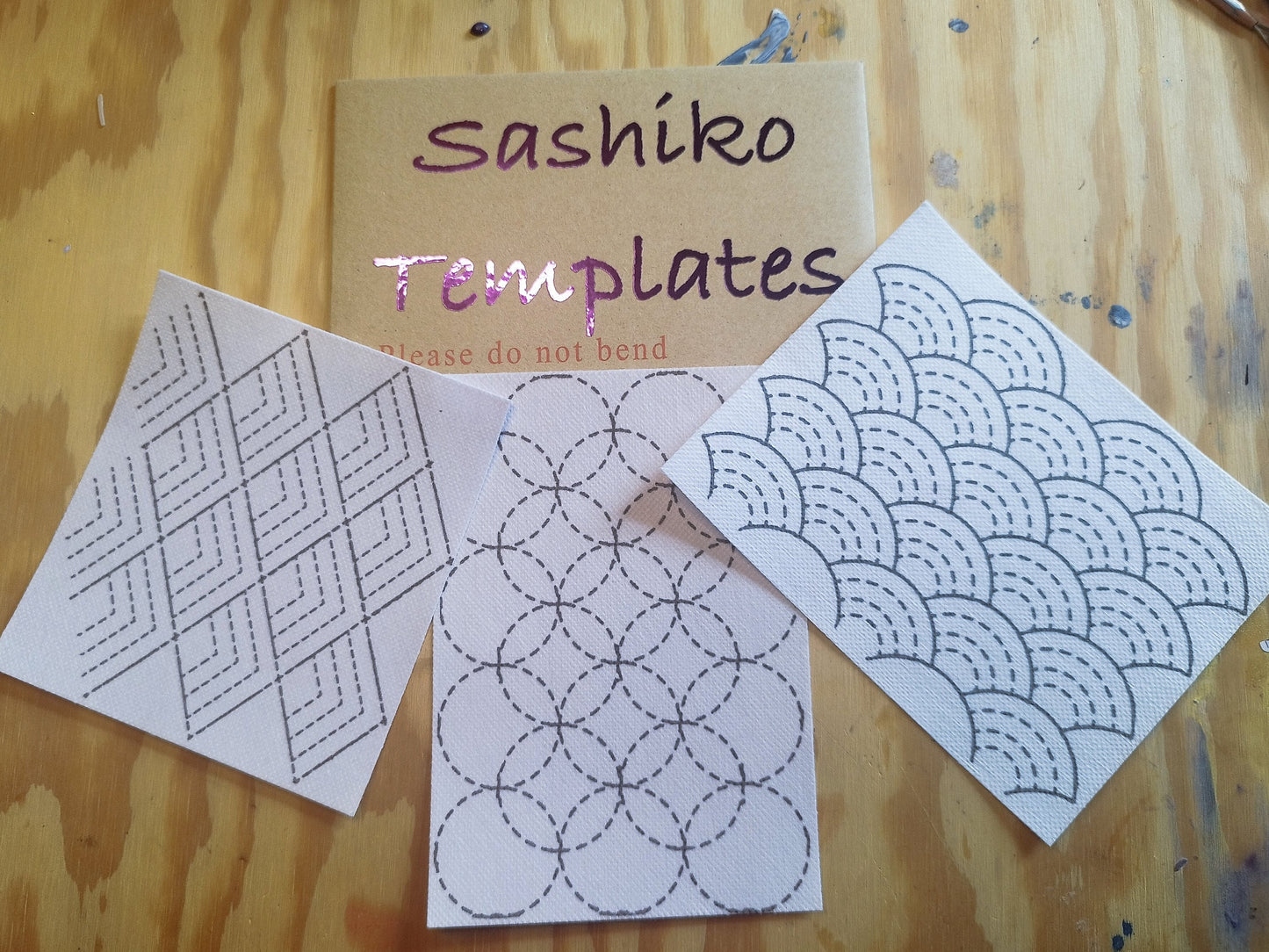 Cardboard hardback envelope with glossy label saying "Sashiko Templates". Beneath is 3 sashiko transfer designs. The design on the left is stacking pyramid shapes. The middle design is overlapping circles. The design on the right is stacking rainbow shapes. On a light wooden background.