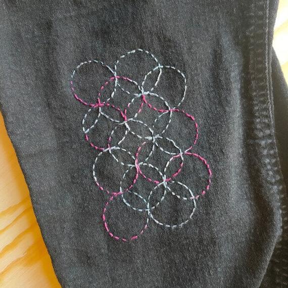 Overlapping Circle Sashiko Design stitched with pink and white thread onto. black denim trouser leg. Some light wood is visible in the background.