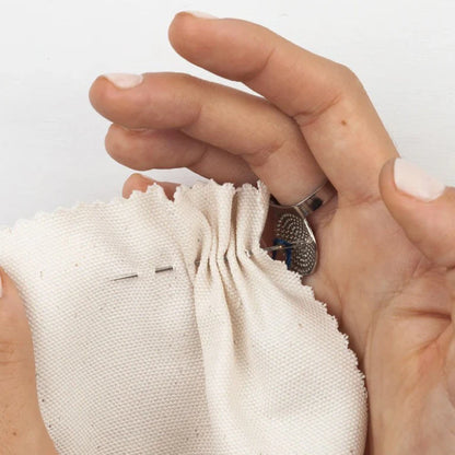 Hand holding a small white fabric pouch with a needle and thread, on a plain background