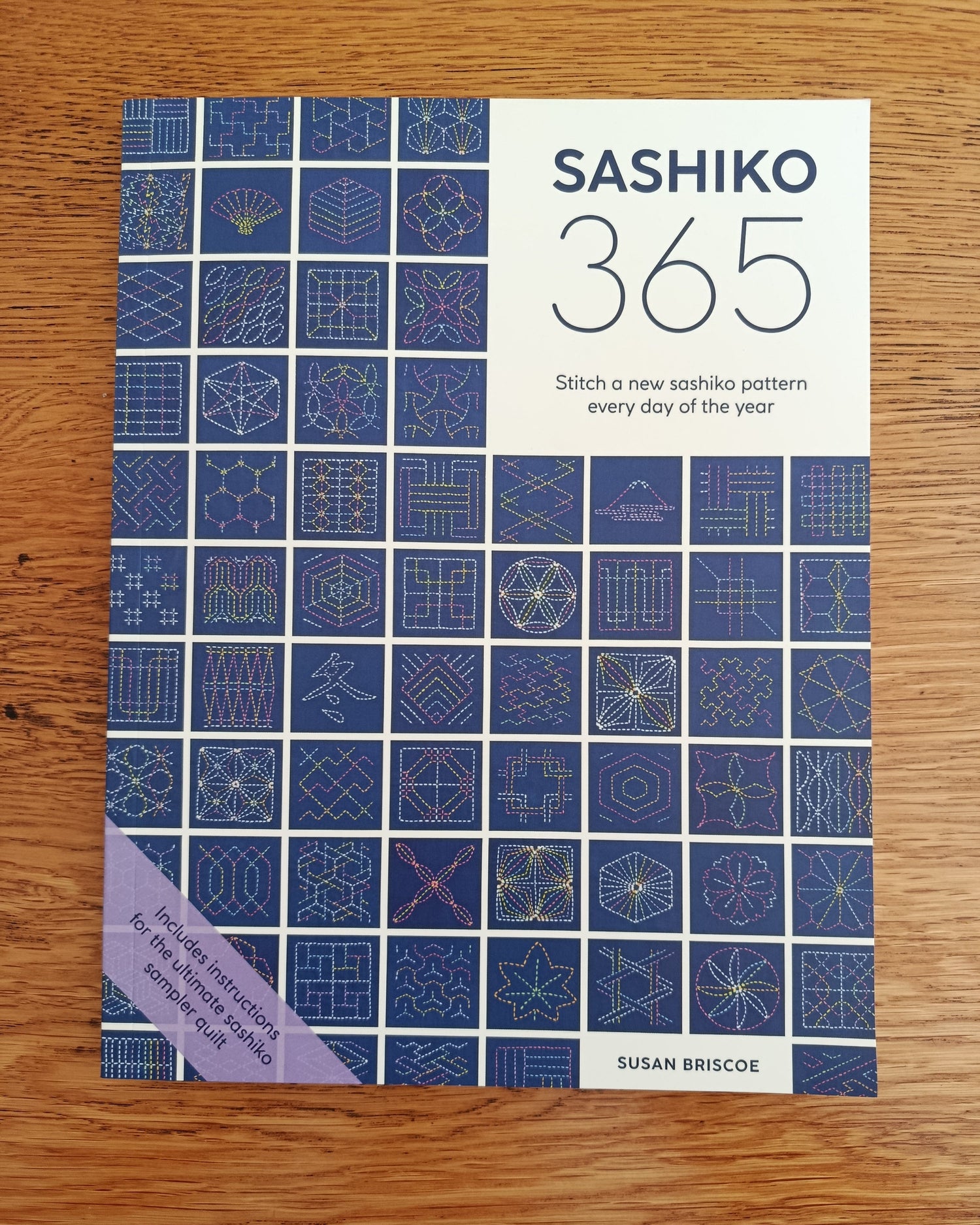 The book cover of 'Sashiko 365' by Susan Briscoe, featuring a blue and white grid pattern of sashiko stitch designs, photographed flat on a wooden surface.
