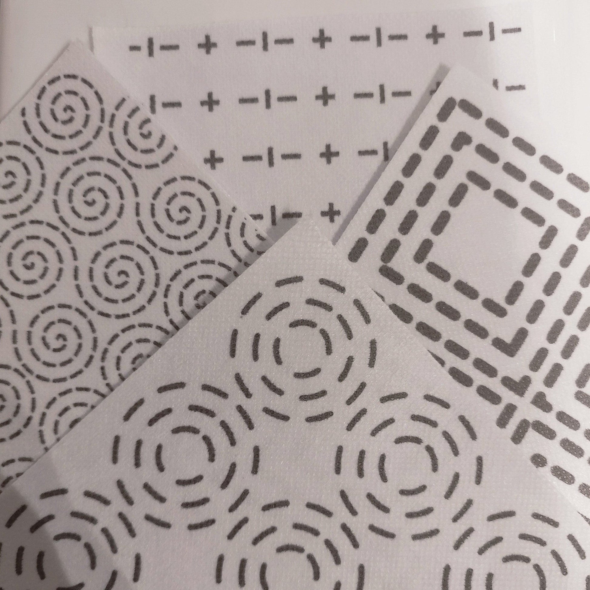 4 Sashiko Transfers. In the foreground is a concentric circles pattern. On the right is a concentric square pattern. At the back is a crosses and dashes sashiko pattern. On the left is a swirls dashed sashiko pattern