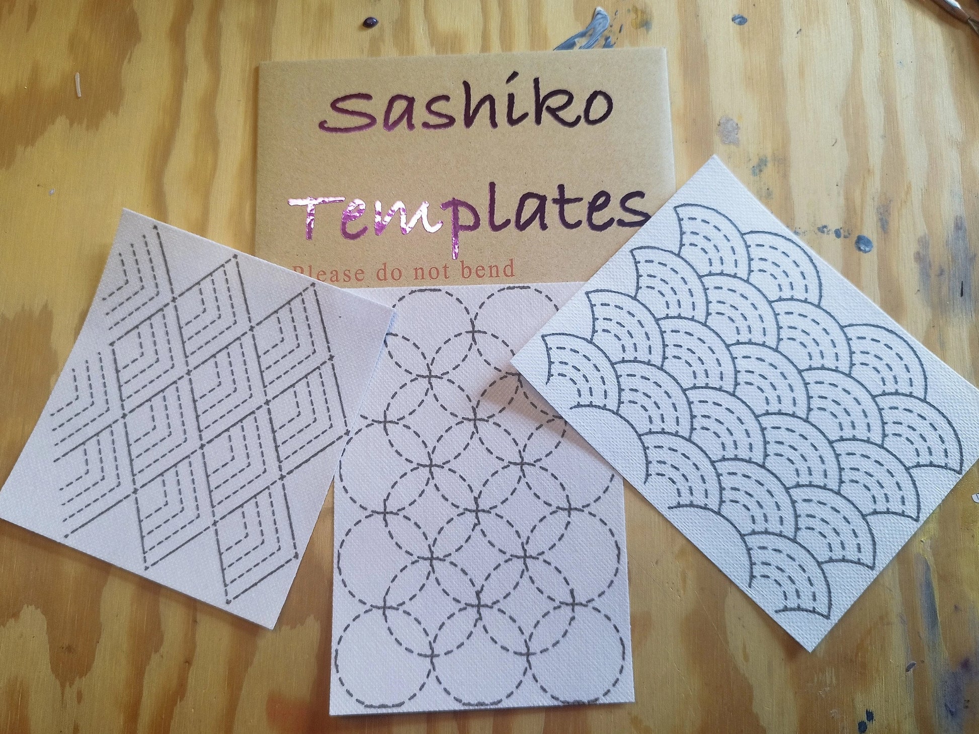 Cardboard hardback envelope with glossy label saying "Sashiko Templates". Beneath is 3 sashiko transfer designs. The design on the left is stacking pyramid shapes. The middle design is overlapping circles. The design on the right is stacking rainbow shapes. On a light wooden background.