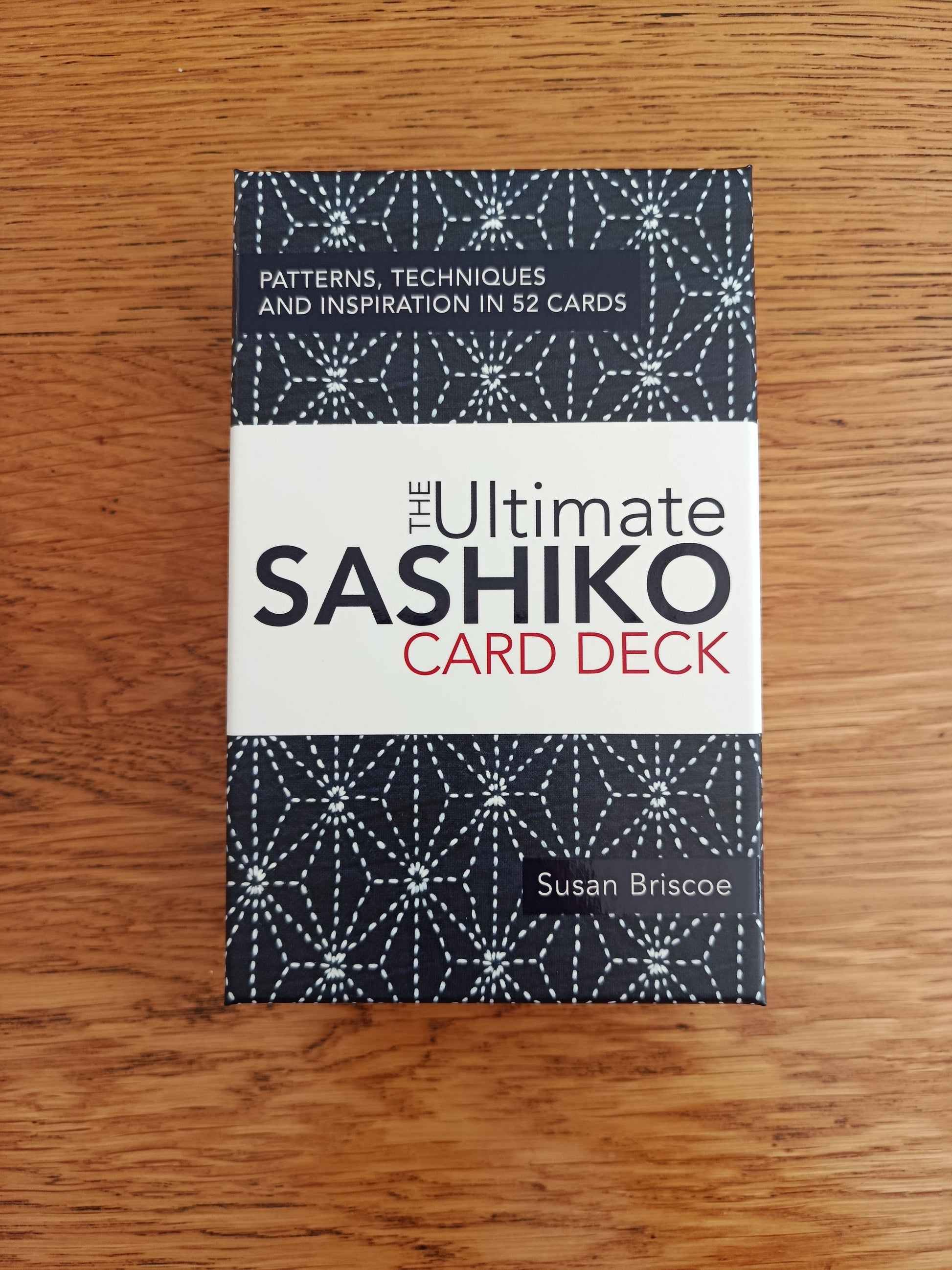 The Ultimate Sashiko Card Deck box by Susan Briscoe, featuring a white center band with black and red text, and patterned dark blue top and bottom sections, placed on a wooden surface.