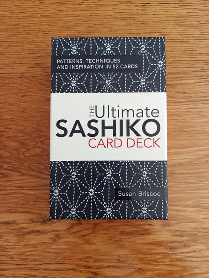 The Ultimate Sashiko Card Deck box by Susan Briscoe, featuring a white center band with black and red text, and patterned dark blue top and bottom sections, placed on a wooden surface.
